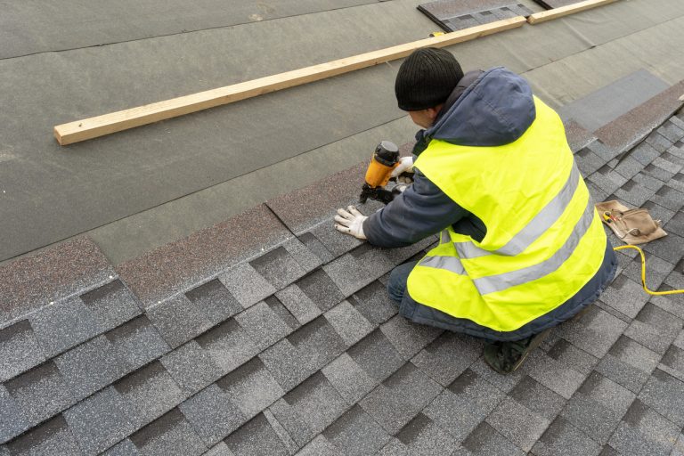 Qualified workman in uniform work wear using air or pneumatic nail gun and installing asphalt or bitumen shingle on top of the new roof under construction residential building