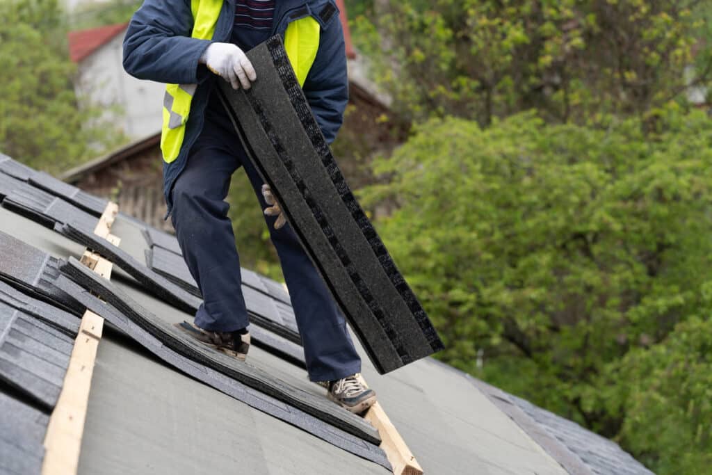 Mature and skilled workman in special protective work wear installing asphalt or bitumen shingle on top of the new roof under construction residential building